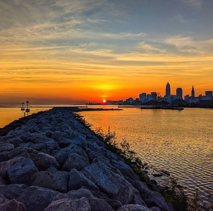 the sun is setting over an ocean with rocks in front of it and buildings in the distance