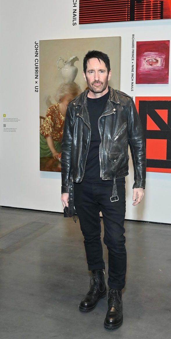 a man in black leather jacket standing next to a wall with art on it and paintings behind him