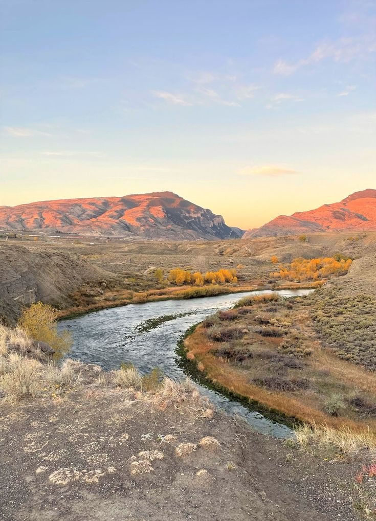 a river running through a dry grass covered field with mountains in the background at sunset