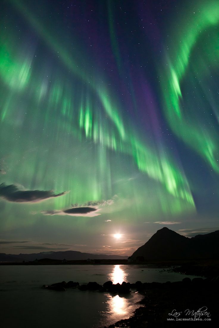 the aurora bore is shining brightly in the night sky over water and rocks, with mountains in the background