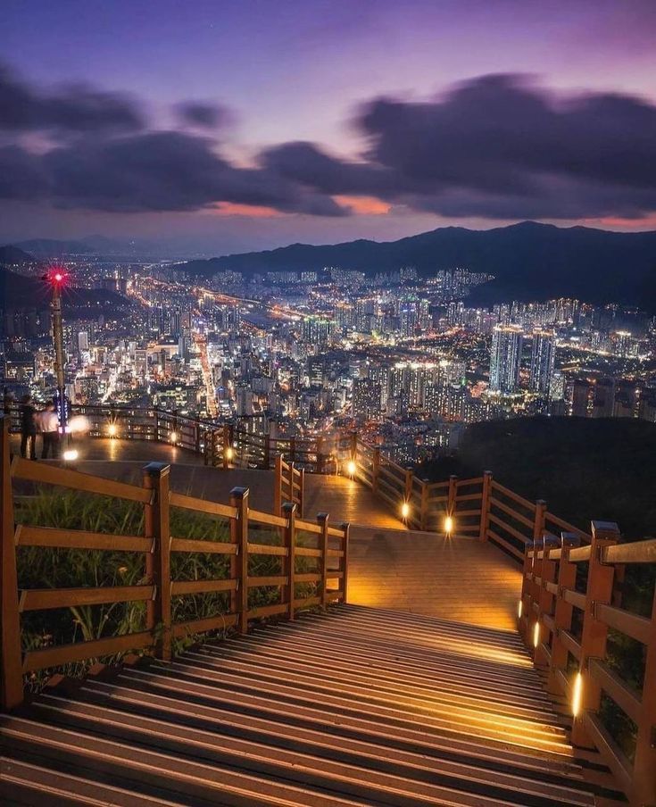 a wooden staircase leading to the top of a hill with city lights in the background