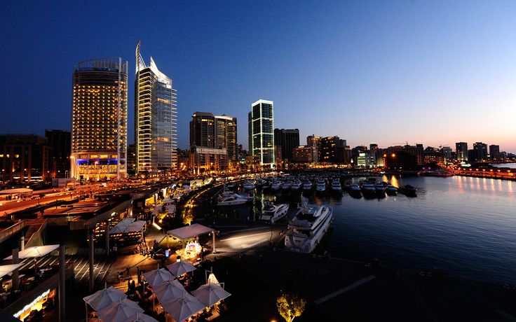 boats are docked in the harbor at night near tall buildings with lights on them and under blue skies