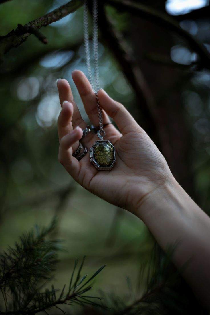 a person's hand holding a necklace in the air with trees behind them and sunlight shining through the branches