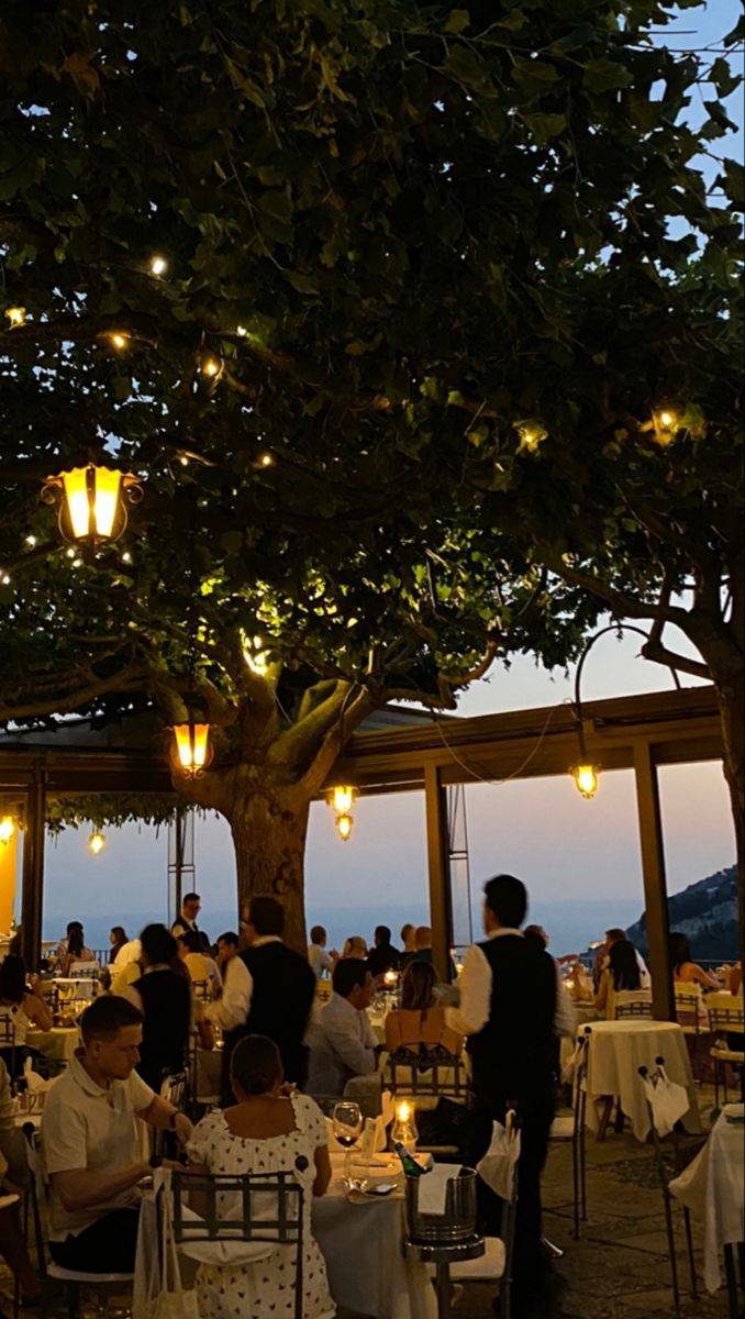 people are sitting at tables under the shade of a tree with lights strung from it