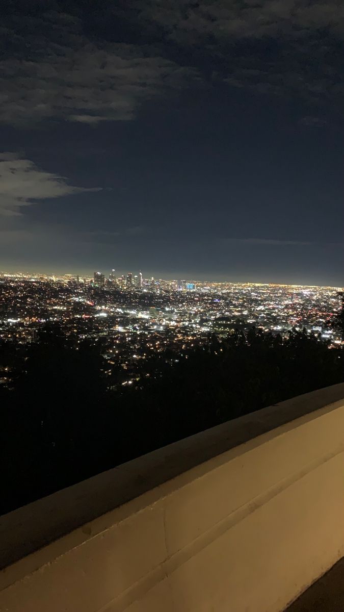 the city lights are lit up in the night sky from atop a hill at an overlook