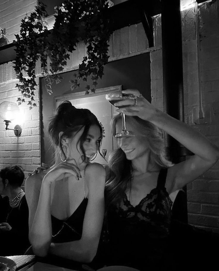 two women sitting at a table with drinks in their hands and one woman holding a wine glass up to her head