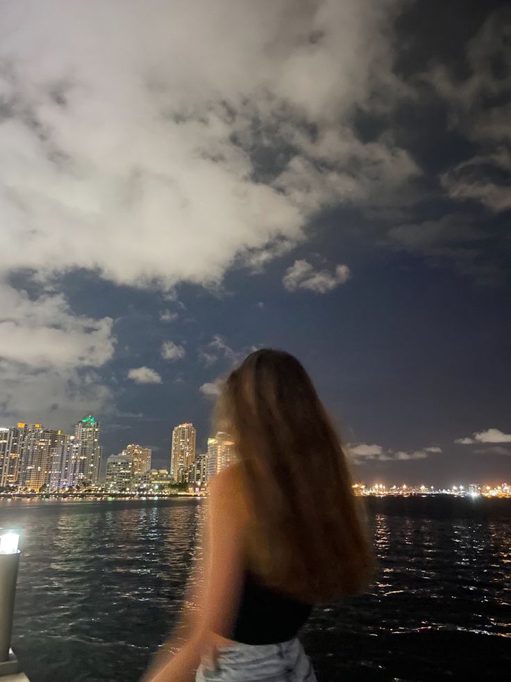 a woman standing on top of a boat looking at the city lights in the distance