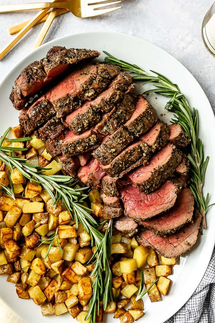 a plate with steak, potatoes and rosemary garnish on it next to silverware