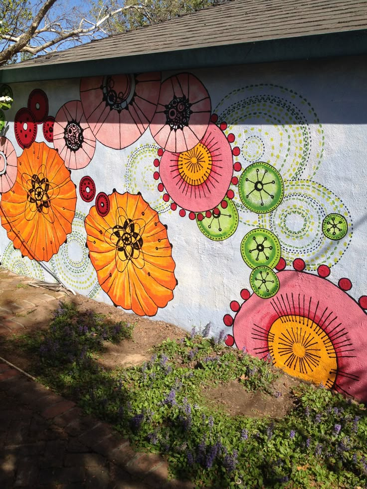 a painted wall with flowers on it in front of a tree and bushes next to a house