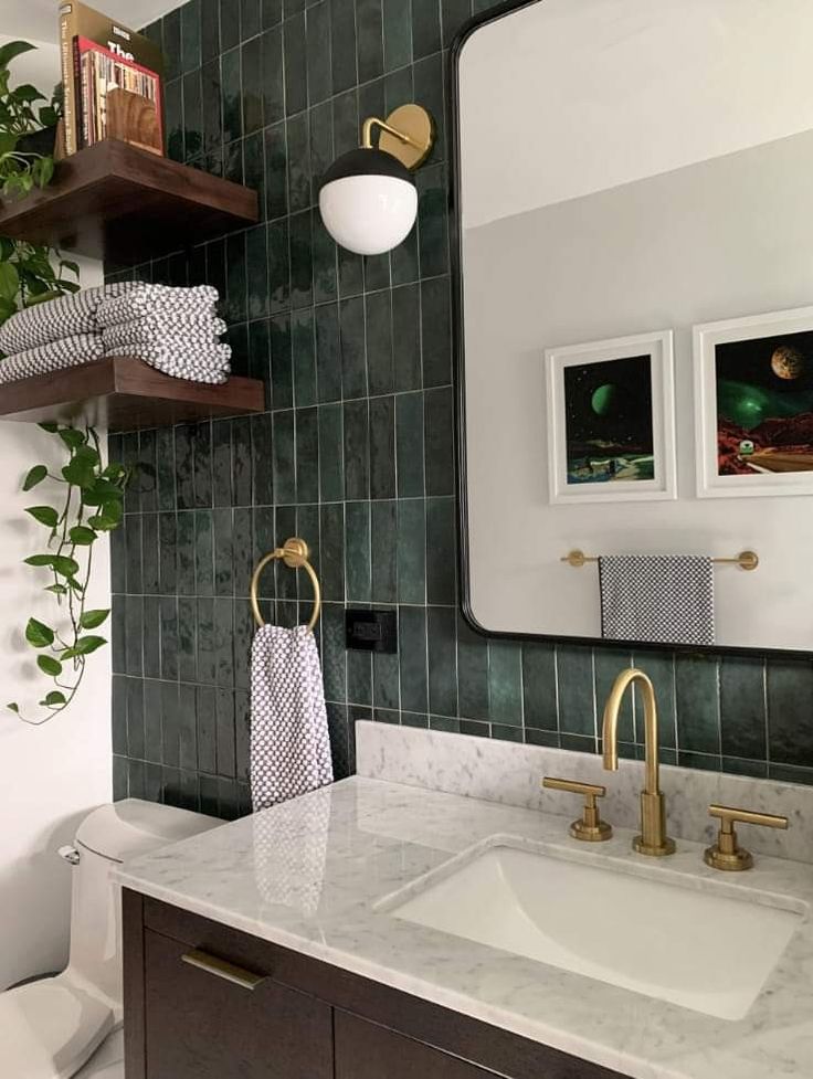 a bathroom with green tiled walls and white counter top, gold faucet, wooden shelving above the sink