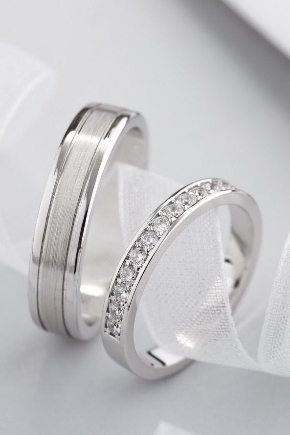 two white gold wedding bands with diamonds on them, sitting next to each other in front of a piece of cloth