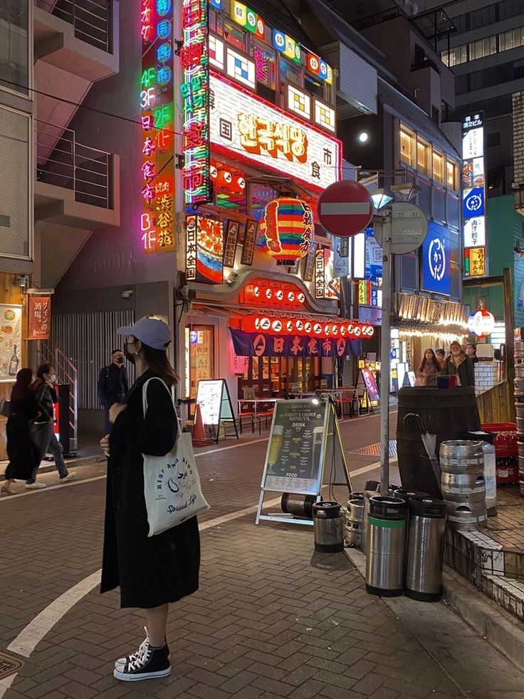 a woman is standing on the sidewalk in front of a building that has neon signs all over it