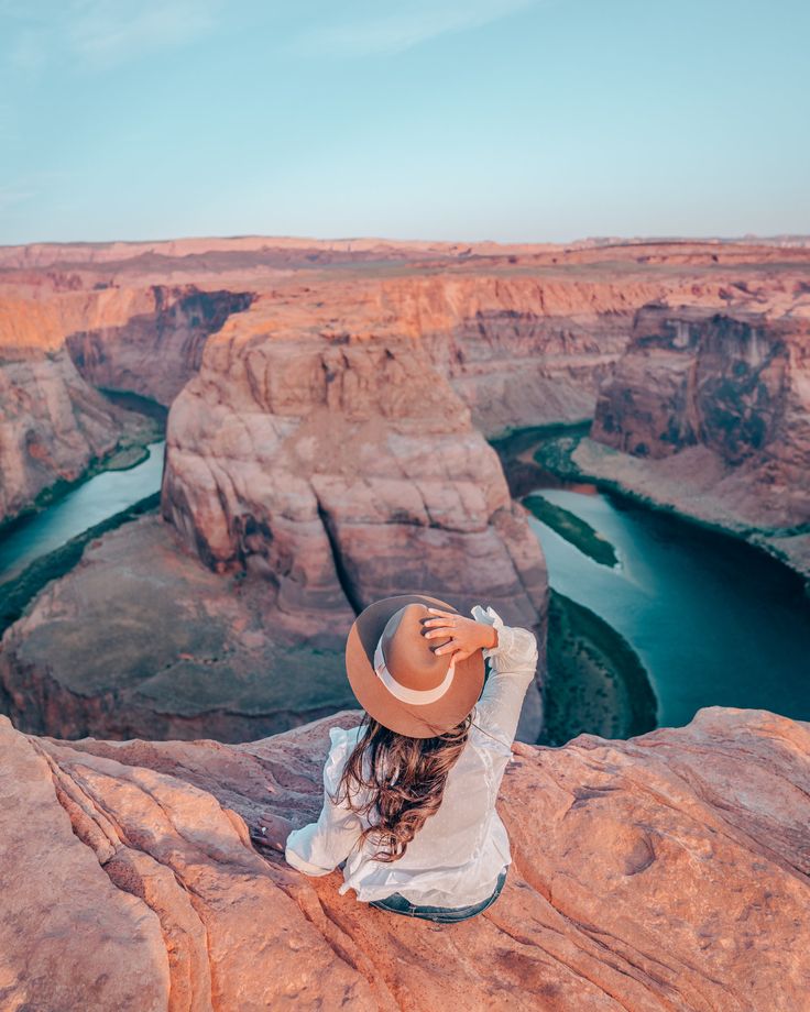 a woman wearing a hat sitting on top of a cliff next to a body of water