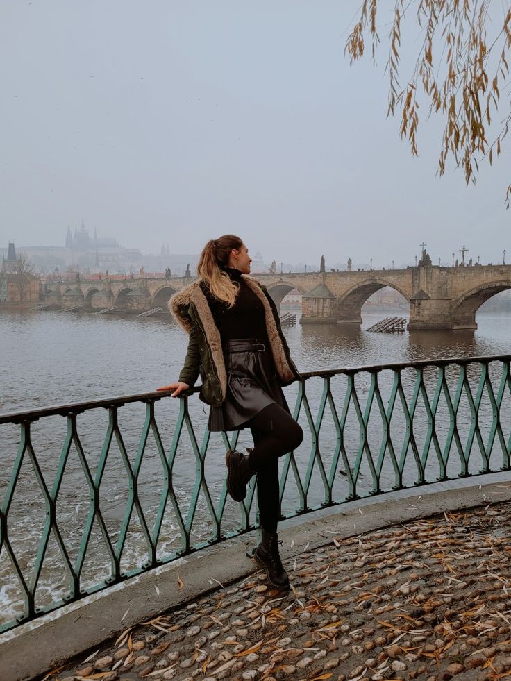 a woman standing on the edge of a bridge next to a body of water with a bridge in the background