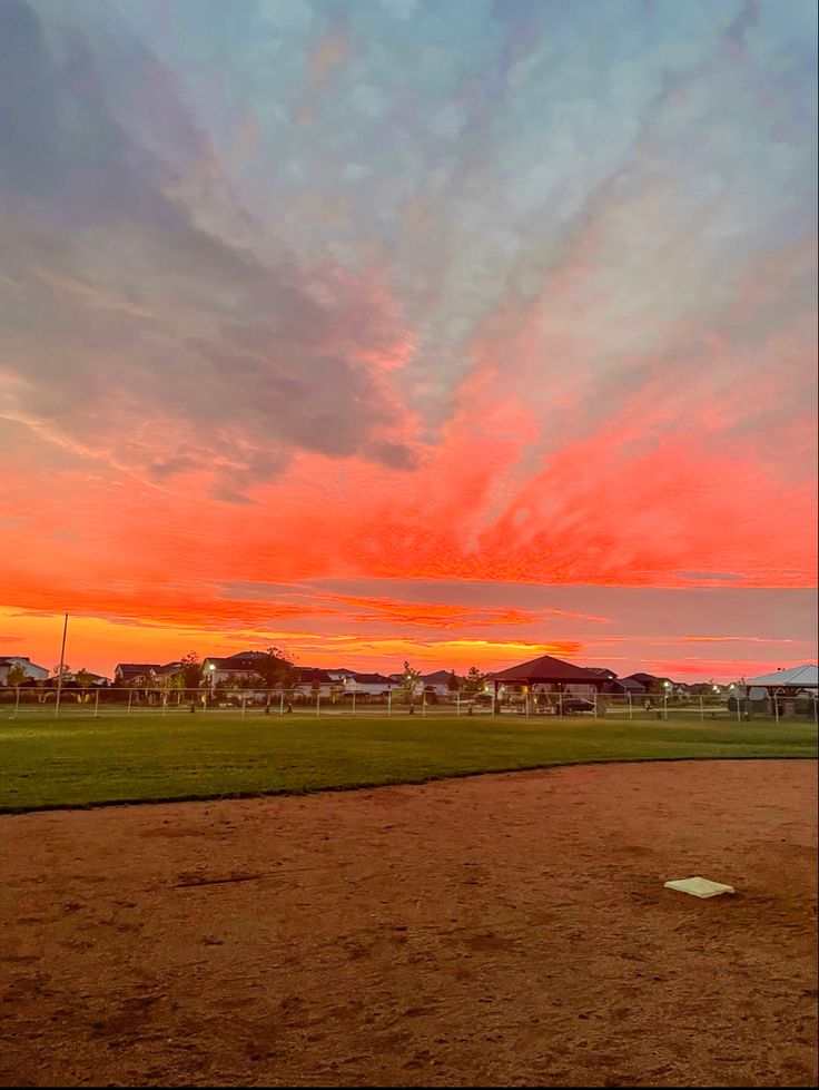 a baseball field with the sun setting in the distance and houses on the other side