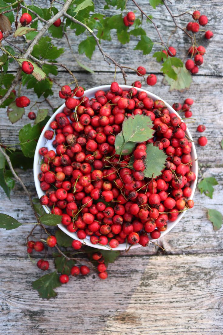 a white bowl filled with red berries on top of a wooden table next to green leaves