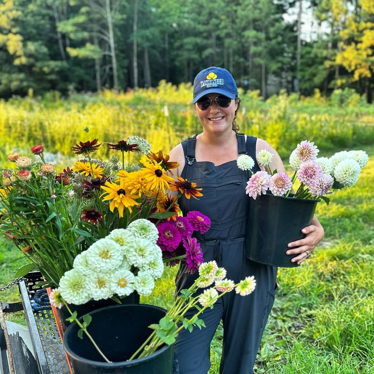 a woman holding two buckets full of flowers in front of some trees and grass