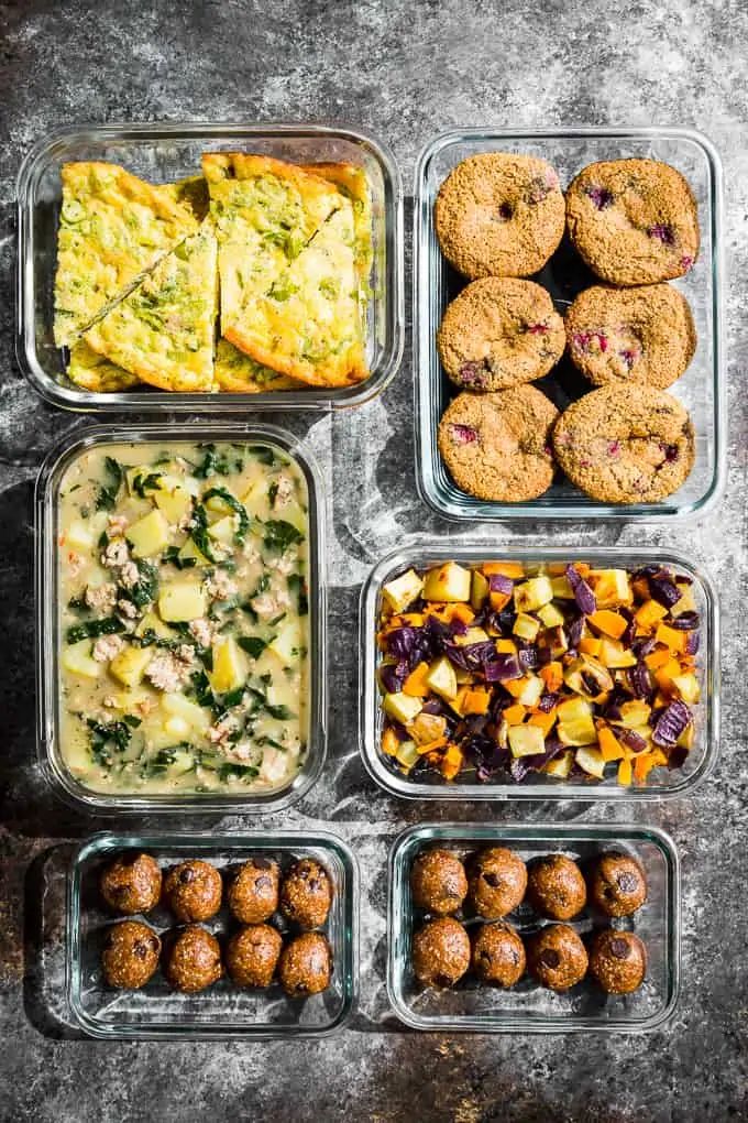 six containers filled with different types of food on top of a gray countertop next to cookies and muffins