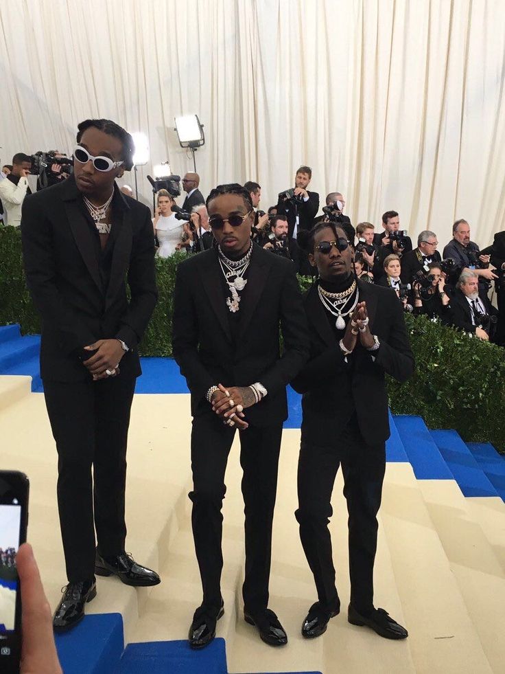 three men in black suits and sunglasses standing on a blue carpet with people taking pictures behind them