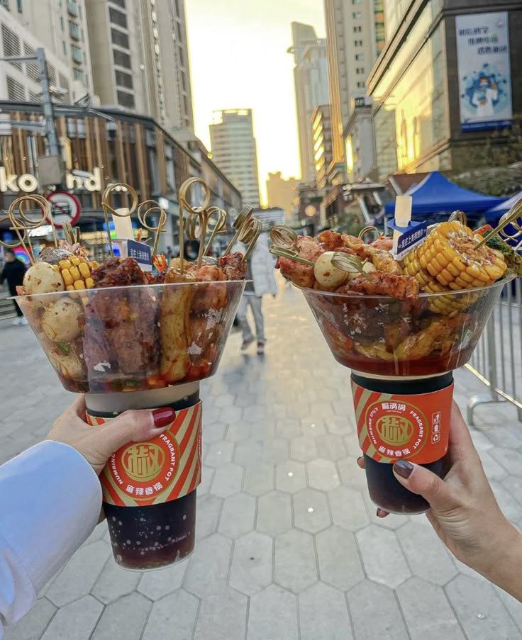 two people holding up cups filled with food on top of a sidewalk in front of tall buildings