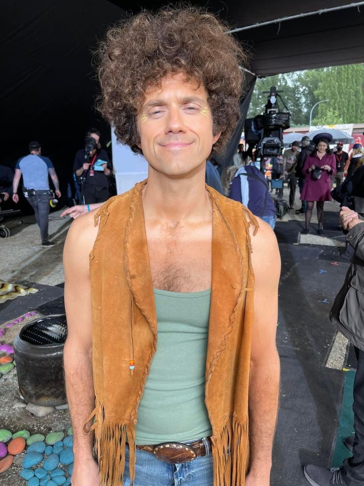 a man with curly hair wearing a brown vest and blue jeans standing in front of a crowd