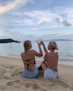 two women sitting on the beach toasting with wine glasses in their hands while looking out at the ocean