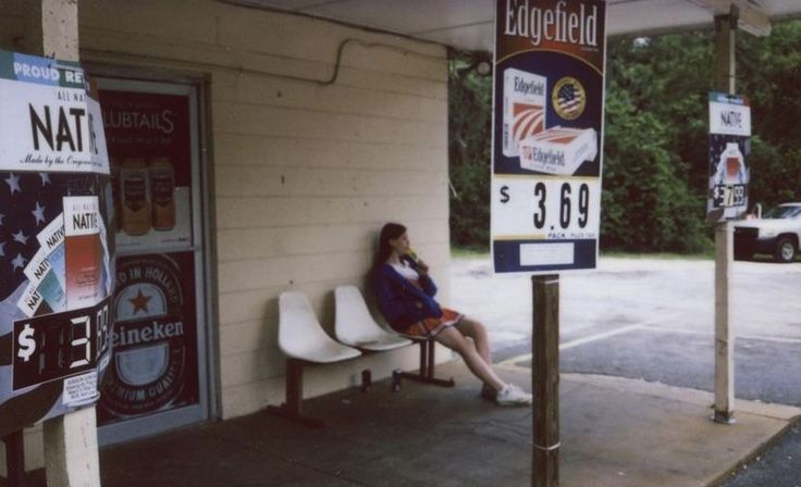 a woman sitting on a bench in front of a gas station with signs and advertisements