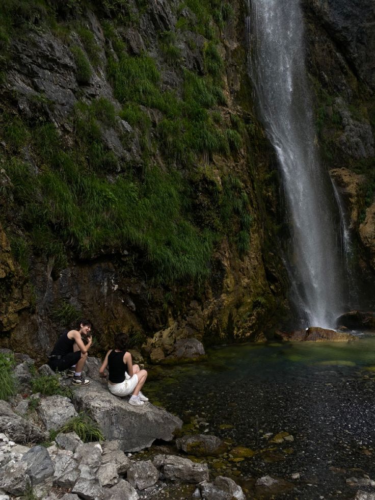 two people sitting on rocks next to a waterfall