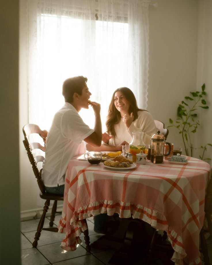 a man and woman sitting at a table in front of a window with food on it