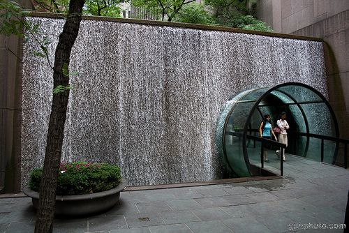 two people standing in front of a waterfall at the entrance to a building with glass walls