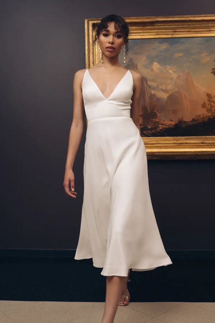 a woman standing in front of a painting wearing a white dress with straps and high heels