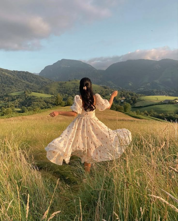 a woman in a white dress is walking through tall grass with mountains in the background