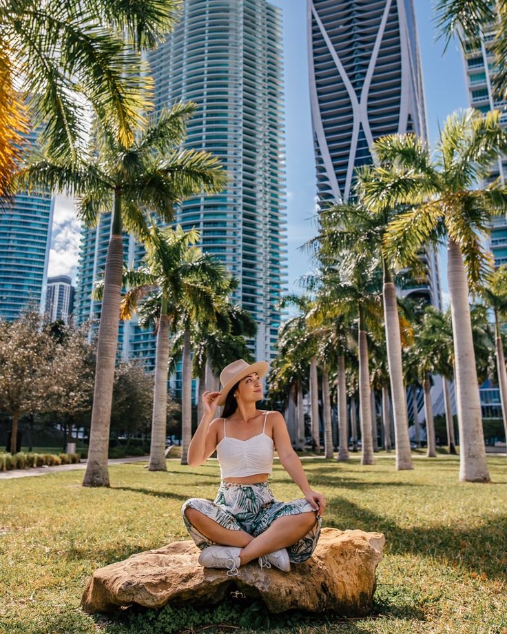 a woman sitting on top of a rock in front of palm trees and tall buildings