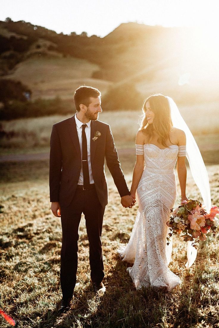 a newly married couple holding hands and walking through the grass in front of mountains at sunset