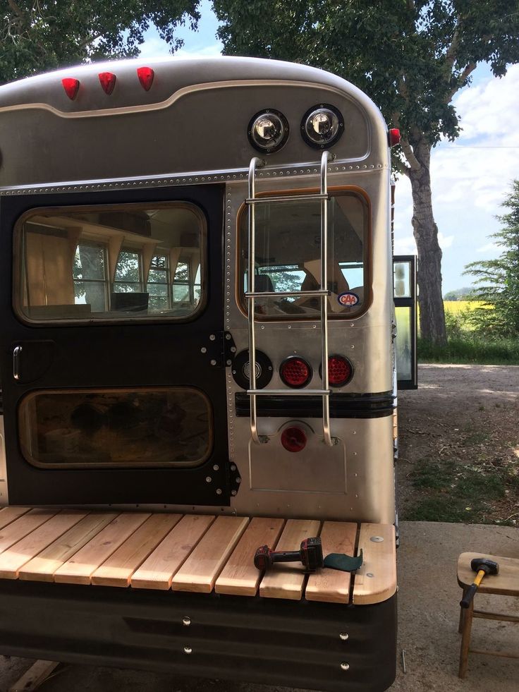 the back end of a silver bus parked on top of a wooden platform next to a tree