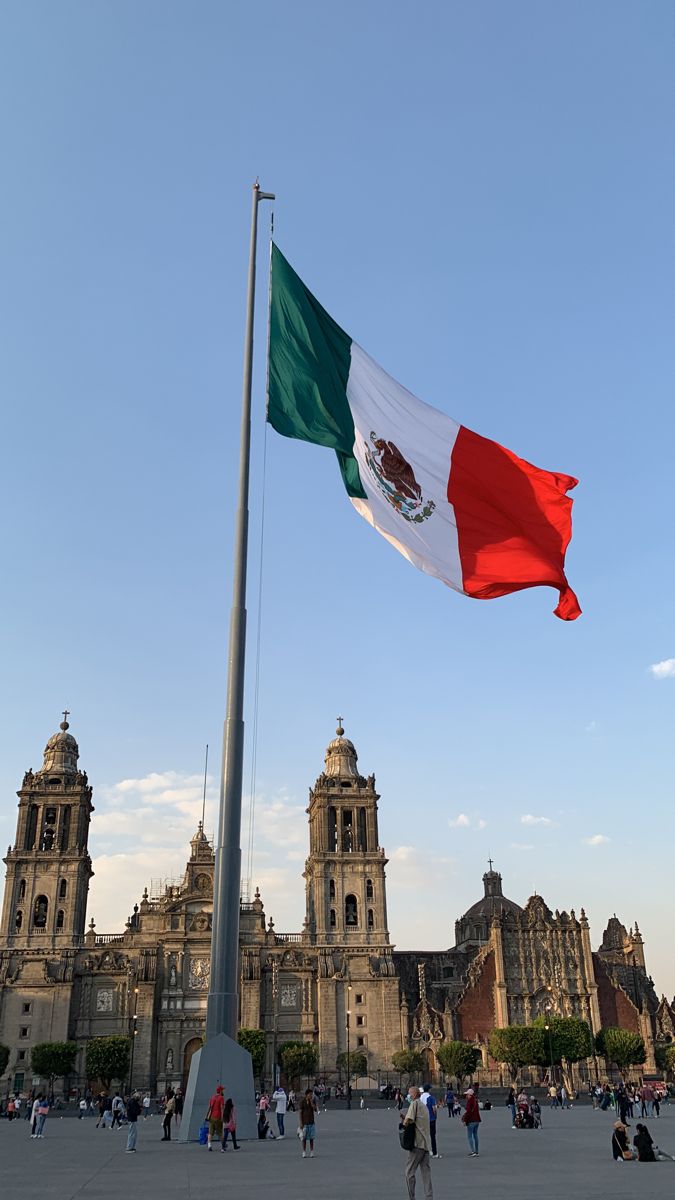 the mexican flag is flying in front of an old building with many people walking around