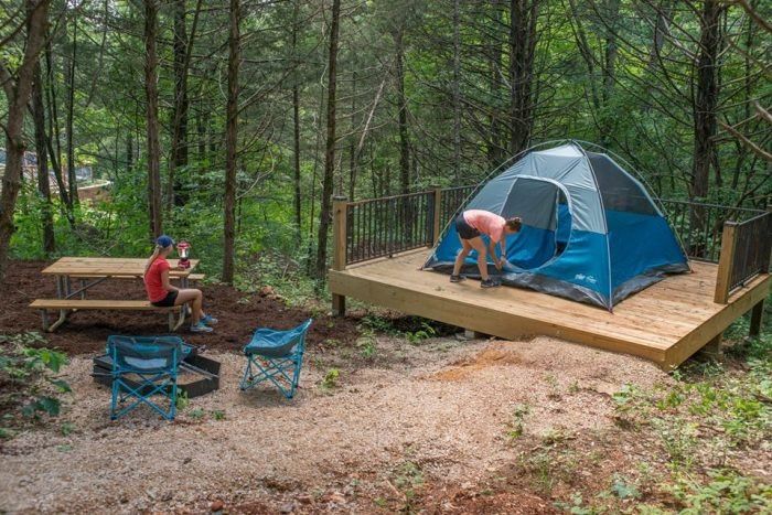 two people setting up a tent in the woods with picnic tables and chairs around it