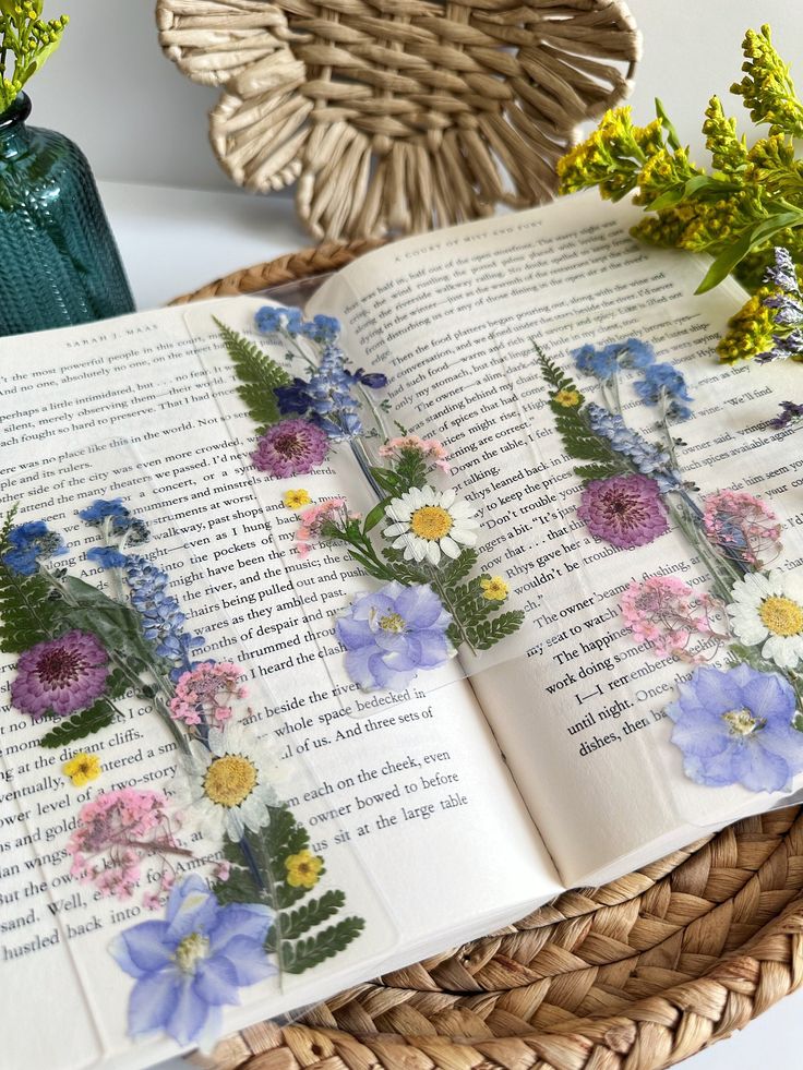 an open book sitting on top of a wicker basket next to flowers and a vase