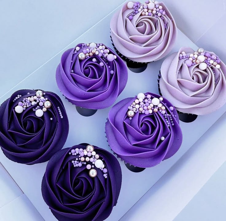 four cupcakes with purple frosting and pearls on top are in a box