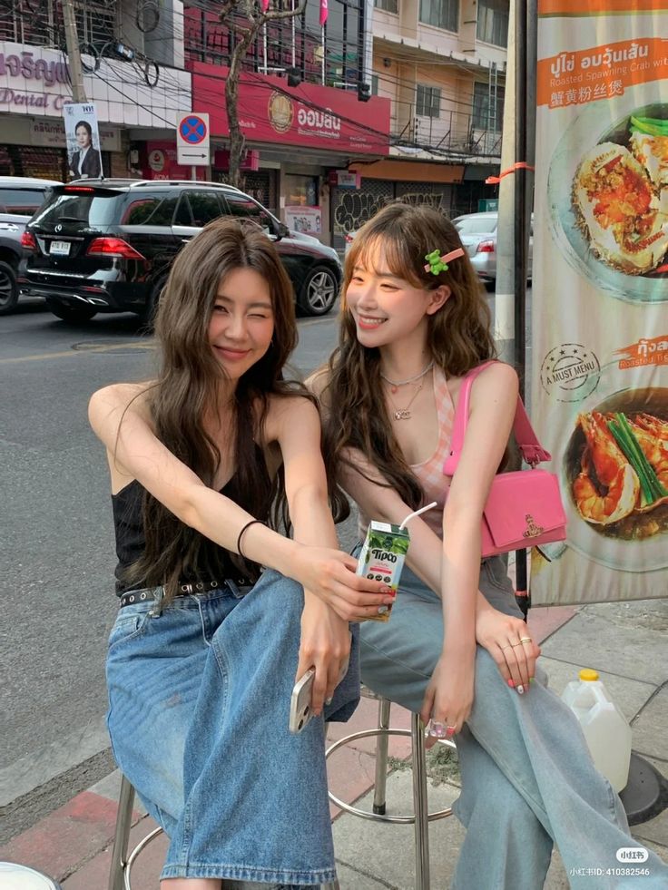 two young women sitting next to each other in front of a food stand on the sidewalk