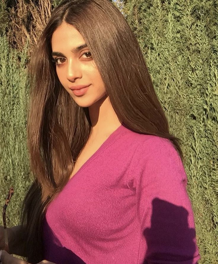 a woman with long brown hair standing in front of some bushes wearing a pink top