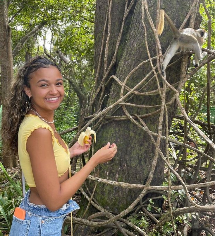 a woman standing next to a tree with a banana in her hand and a monkey hanging from the tree