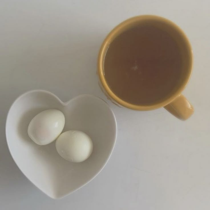 two eggs in a heart shaped bowl next to a cup of tea