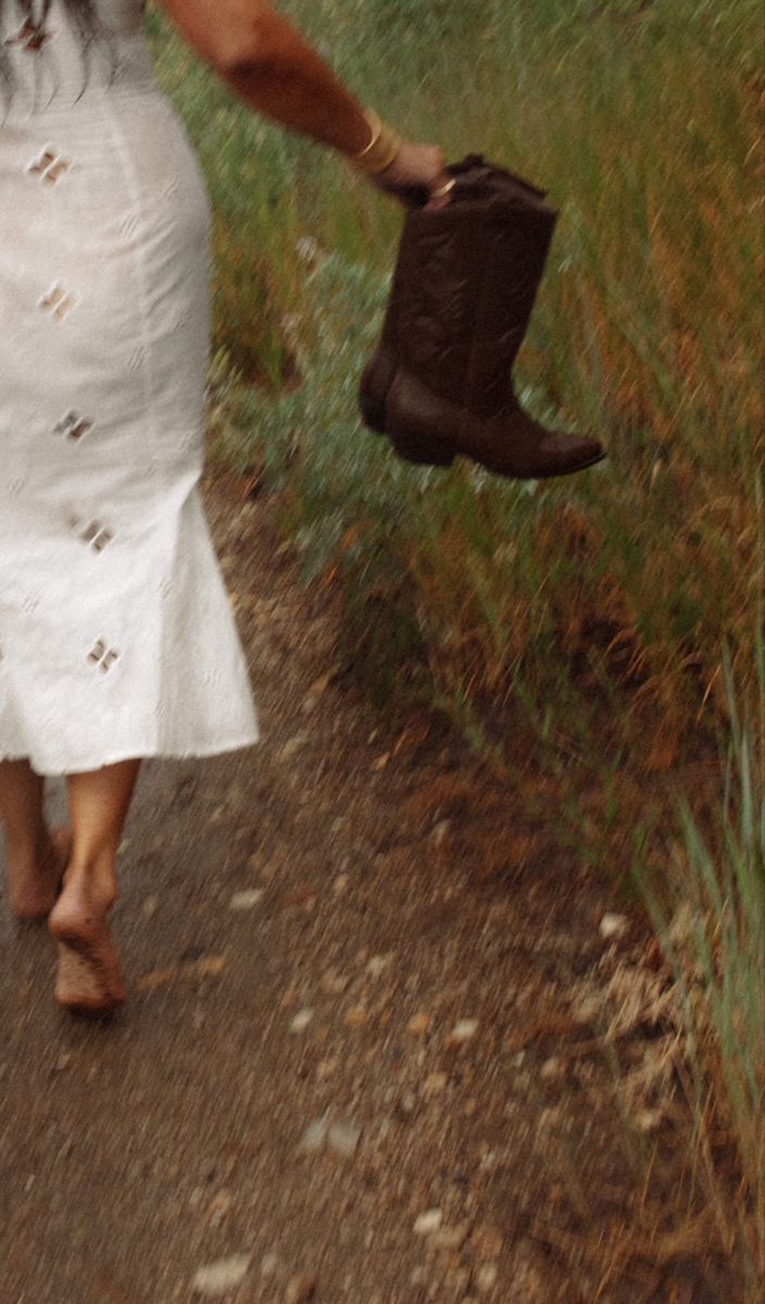 a woman walking down a dirt road holding a boot in her hand and wearing a white dress