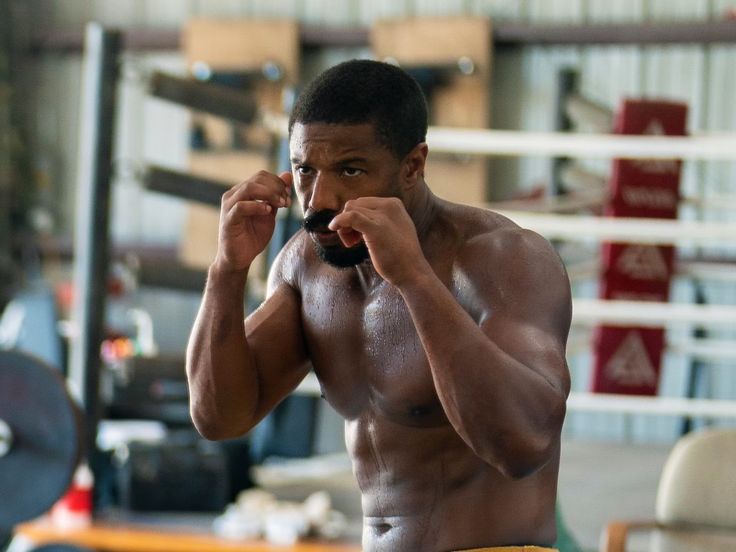 a shirtless man holding a black object in his right hand and looking at the camera