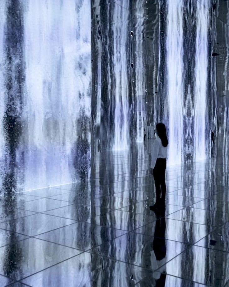 a woman standing in front of a waterfall with her reflection on the ground and another person looking at it