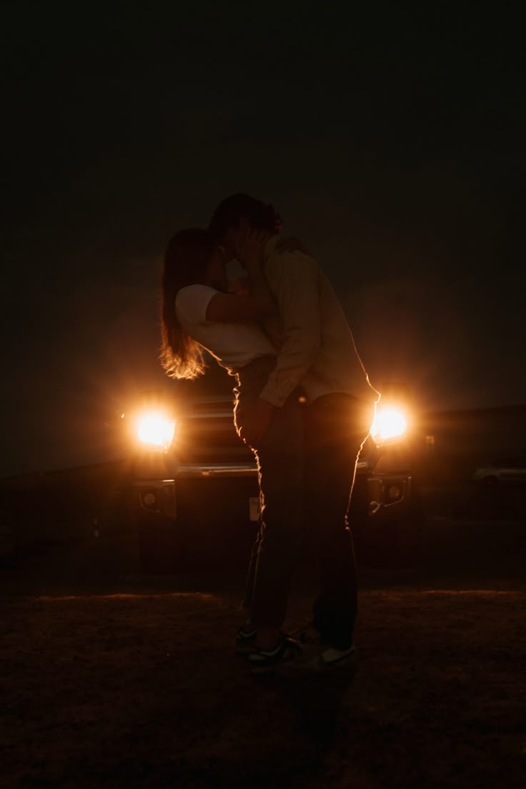 a man and woman kissing in front of a truck at night with the headlights on