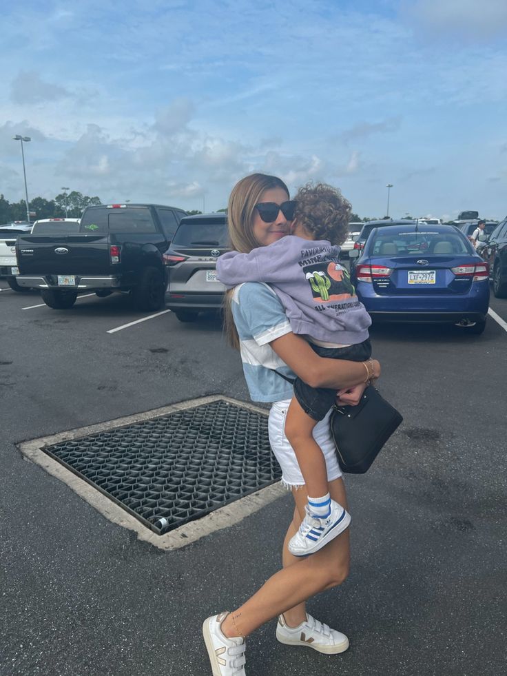 a woman carrying a child in her arms while walking through a parking lot with other cars