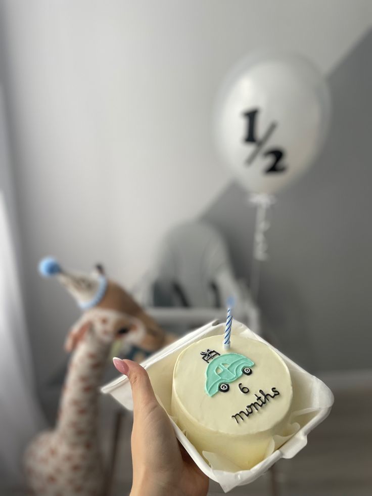 a person holding a birthday cake in front of a small giraffe and balloon