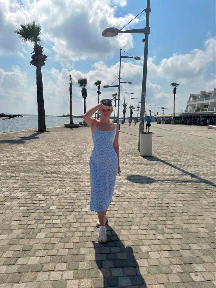 a woman in a blue dress standing on a brick road next to the ocean and palm trees